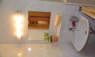 A bright bathroom with white tiles and a small sink. A potted plant and fresh towels are on the shelf.