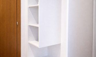 A modern wardrobe made of white wood with several shelves and a clothes rod. The floor is covered with a red carpet and a wooden door is visible in the background.
