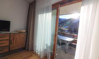 A bright room with a balcony door, through which sunlight streams. Outside is a balcony overlooking the mountains.