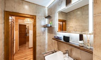 A modern bathroom with a sink and a large mirror. The floor is made of wood, and there are additional furniture pieces in the background.