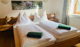 A cozy bedroom with two beds, surrounded by wood and bright light. Green towels are placed on the beds and a decorative picture hangs on the wall.