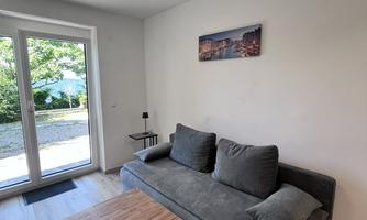 A modern living room with a gray sofa and a wooden table. Large windows offer a view of the outdoor area.