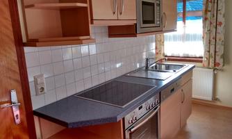 A modern kitchen with wooden cabinets and a cooktop. It also includes a sink and an oven, with plenty of daylight through the window.