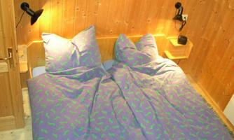 A cozy bedroom with a double bed and colorful duvets. The walls are made of wood, and there are two bedside lamps.