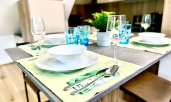 A modern set table in a cozy kitchen. There are white plates, glasses, and cutlery, surrounded by green accents.