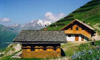 A traditional alpine hut in a green mountain landscape. In the background, snow-capped mountains and a blue sky are visible.
