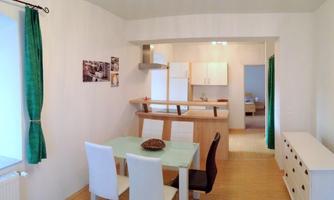 A bright room with a dining table and four chairs. In the background, a modern kitchen is visible.