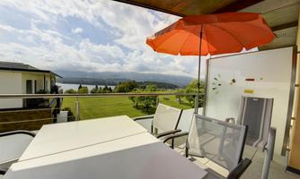 A balcony with a table and chairs. A sun umbrella protects from the sun while enjoying the view of the lake and the green landscape.