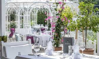 An elegant restaurant with white tables and decorative flowers. In the background, a pavilion is visible, surrounded by green plants.