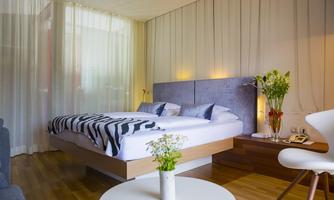 A modern bedroom with a large bed and elegant bed linens. The room is bright and friendly with plants and a table in the foreground.