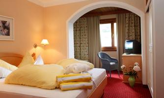 A cozy bedroom with a large bed, yellow pillows, and a comfortable seating area. The room has bright walls and a window with curtains.
