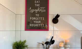 A modern kitchen with an inspiring mural that includes the invitation to eat spaghetti. On the countertop are a plant, dishes, and candles.