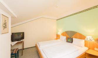 A cozy room with two single beds and a television. The walls are in soft colors, and there is a table lamp on a nightstand.