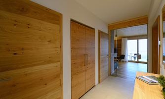 A modern hallway with wooden doors and bright walls. Natural light streams through the large windows into the adjacent room.