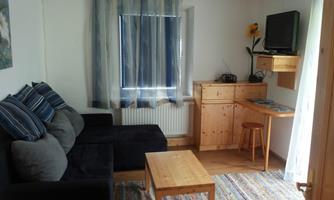 A cozy room with a black sofa, a wooden table, and a desk. The walls are light, and there are curtains with blue stripes.