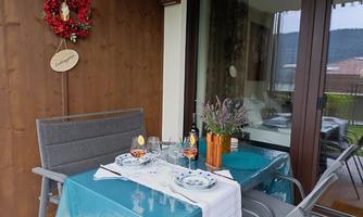 A cozy balcony with a beautifully set table, decorated with glasses and a bouquet of lavender. In the background, there is a wooden panel and a festive wreath decoration.