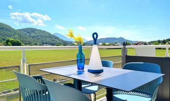 A sunny terrace with a table and blue chairs. In the background, green mountains and a clear sky can be seen.