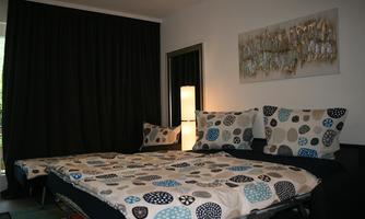 A cozy bedroom with a large bed, different pillows, and a bedside table. The walls are dark, and there is an artistic picture on the wall.