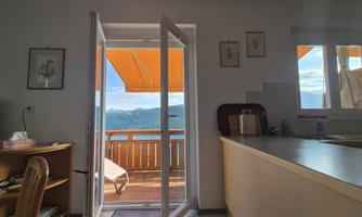 A bright interior view with open doors leading to a balcony with a beautiful view. Outside, there are orange awnings and a picturesque landscape.