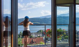 A woman stands on a balcony looking out at a picturesque lake and the surrounding mountains. The sky is clear and the landscape is green and inviting.