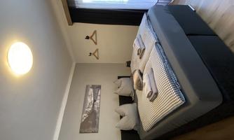 A modern bedroom with a large bed and a bright window. The walls are gray, and there is a painting as well as some clothes hooks.