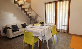 A modern living room with a dining table and colorful chairs. In the background, a staircase and large windows with curtains are visible.