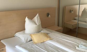 A bright bedroom with a double bed and a large mirror. An artwork hangs on the wall, and on the bed are pillows and blankets.
