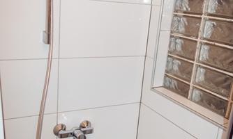 A modern shower cabin with white tiles and a glass wall. The shower is equipped with a handheld showerhead and a modern faucet set.