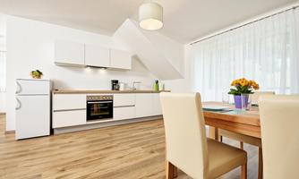 A modern kitchen with white cabinets and a wooden floor. The dining area is bright and inviting, with a table and a floral decoration.