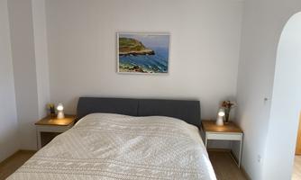 A cozy bedroom with a large bed and nightstands.  
On the wall hangs a picture of a coastal landscape.