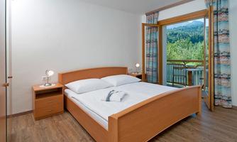 A cozy bedroom with a double bed and wooden furniture. Large windows offer a view of the green landscape and the balcony.