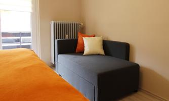 A modern room with a gray sofa and orange cushions. In the background, a bed with an orange blanket and a radiator is visible.