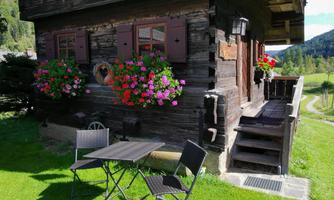 A cozy wooden house with blooming flower boxes at the windows. In front of the house, there is a table and chairs in a green meadow.