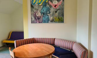 A cozy seating area with a round table and an upholstered bench. A colorful piece of art titled "007" hangs on the wall.