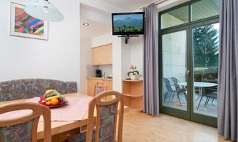 An inviting dining area with a wooden table and colorful chairs. Large windows offer a view of a garden and the television hangs on the wall.