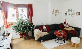 A festively decorated living space with a Christmas tree and red curtains. The cozy couch is adorned with decorative pillows and a small table.