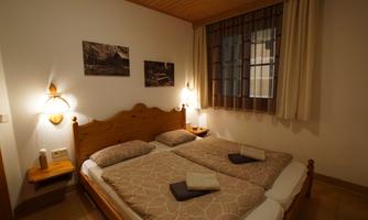 A cozy double room with two beds, decorated in warm wood tones. The walls are adorned with pictures and the room has windows with curtains.