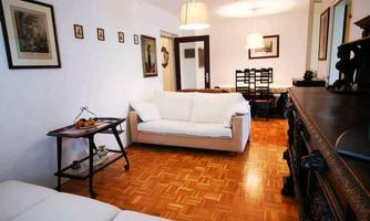 A cozy living room with a light wooden floor and modern furniture. Various pictures hang on the walls, and there is ample light from the ceiling lamps.