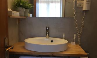 A modern sink on a wooden shelf in a bathroom. In the background, plants and a window can be seen.