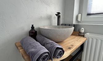 A modern stone sink on a wooden table. Next to it are two folded towels and a bottle of care product.