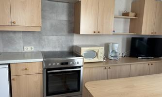 A modern kitchen with wooden cabinets and a bright countertop. Visible are an oven, a microwave, and a television.