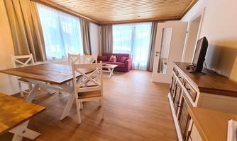 A bright living room with wooden floors and large windows. There is a dining area with a table and chairs as well as a cozy seating corner with a pink sofa.
