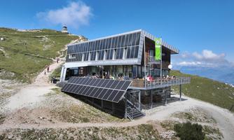 A modern mountain house with large windows and a terrace. Surrounded by green meadows and a clear blue sky.