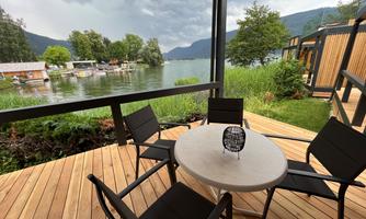 A cozy terrace with a round table and chairs that overlooks a tranquil lake. In the background, green trees and some boats are visible.