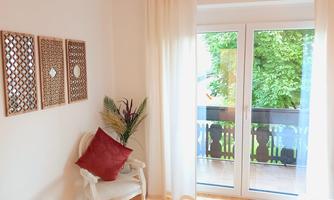 A bright room with a comfy chair and a red cushion.  
Large windows lead to a balcony with a view of greenery.