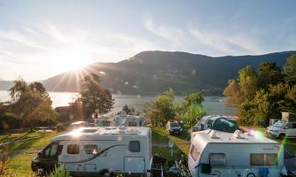 A campsite by the water with motorhomes and a beautiful view of the mountains. The sun shines over the landscape, creating a cozy atmosphere.