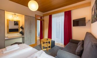 A modern room with a double bed and a gray sofa. There is a desk and a television, as well as large windows with red curtains.