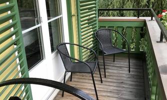 A balcony with two black chairs and green window shutters. In the background, plants and a quiet residential area are visible.