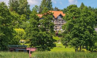 A picturesque house lies among tall trees by the shore of a clear lake. The surroundings are green and inviting, with a beautiful view of nature.