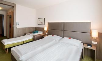 A modern hotel room with two single beds and an extra bed. The walls are light and the lighting is pleasant and cozy.
